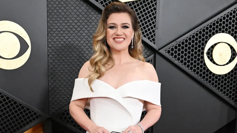 Kelly Clarkson smiling on red carpet in white off-shoulder gown